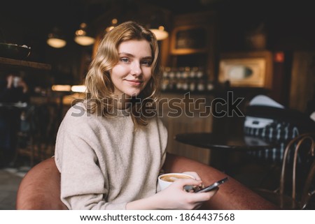 Content woman in casual sweater sitting in comfortable armchair with cup of hot coffee and messaging on smartphone while looking at camera