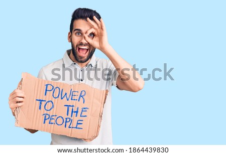 Young handsome man with beard holding power to the people cardboard banner smiling happy doing ok sign with hand on eye looking through fingers 