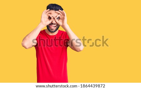 Young handsome man with beard wearing casual t-shirt doing ok gesture like binoculars sticking tongue out, eyes looking through fingers. crazy expression. 