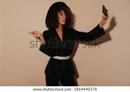 Cool young woman with fluffy hairstyle in shiny modern sweater and hat making selfie and showing tongue on isolated backdrop..