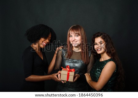 Indoor portrait of three happy bestfriends sisters women, ready for holiday party, holding bright gifts and presents, wearing bright dresses on black background
