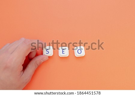 A hand is placing the letter "S" to form the acronym "SEO", for Search engine optimization, over an orange background. Web search engines and its importance concepts