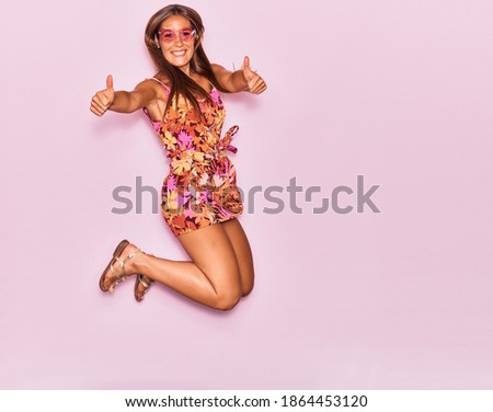 Young beautiful hispanic woman wearing casual clothes and sunglasses with heart shape smiling happy. Jumping with smile on face doing ok sign with thumbs up over isolated pink background.