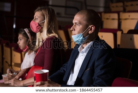 Portrait of man wearing medical face mask sitting in cinema with his family, watching movie. Entertainment concept in coronavirus pandemic