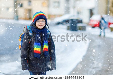 Little school kid boy of elementary class walking to school during snowfall. Happy healthy child with glasses having fun and playing with snow. With backpack or satchel in colorful winter clothes.