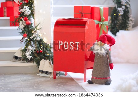 
A gnome is standing next to a red mailbox. White skates lie in the background snow, the veranda is white. The verandahs are decorated with a Christmas tree and garlands