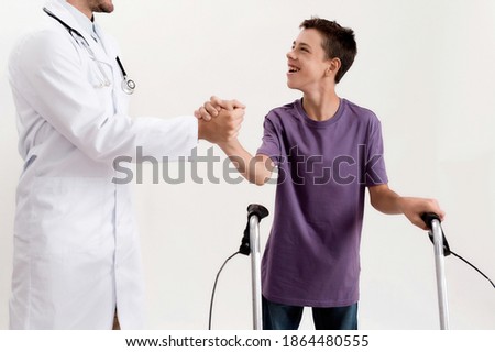 Cropped shot of male doctor shaking hands with cheerful disabled boy with cerebral palsy, taking steps using his walker isolated over white background. Children with disabilities and special needs Royalty-Free Stock Photo #1864480555