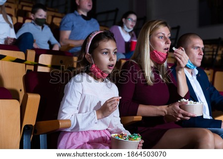Portrait of cute preteen girl visiting cinema with parents during coronavirus pandemic. Girl wearing protective mask eating popcorn and absorbedly watching movie