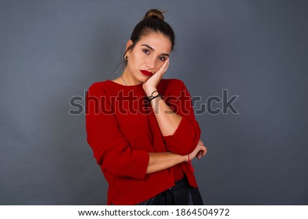 Very bored Young beautiful Caucasian woman wearing red sweater against gray background  holding hand on cheek while support it with another crossed hand, looking tired and sick,