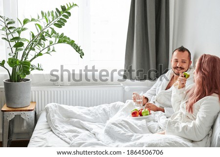 side view on cheerful couple have breakfast on bed at weekend, after waking up. they lie together on bed in bathrobes, eat healthy food