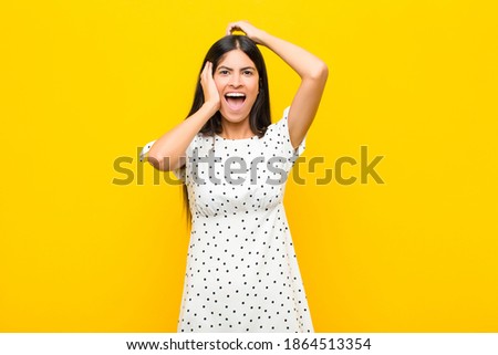 young pretty latin woman raising hands to head, open-mouthed, feeling extremely lucky, surprised, excited and happy against flat wall