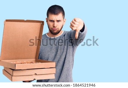 Young handsome man holding delivery italian pizza boxes with angry face, negative sign showing dislike with thumbs down, rejection concept 