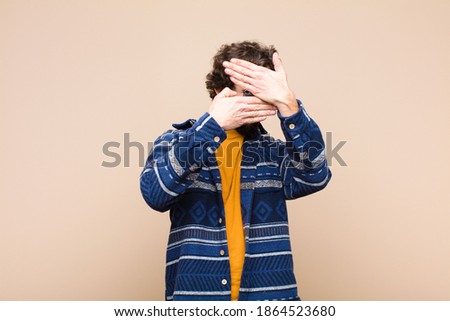 young crazy cool man covering face with both hands saying no to the camera! refusing pictures or forbidding photos against flat wall