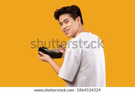Finance, Investment And Money Saving. Portrait of excited asian young man holding and showing wallet full of dollars, free space, isolated over orange studio background. Greedy guy got salary Royalty-Free Stock Photo #1864534324