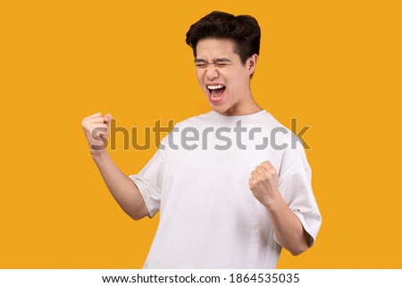 Yeah, I Did It. Portrait of overjoyed asian guy screaming with joy and clenched fists raised up in the air, isolated over orange background. Excited young man cheering and yelling, celebrating victory Royalty-Free Stock Photo #1864535035