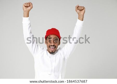 Excited joyful young african american man 20s years old in basic streetwear hoodie standing doing winner gesture clenching fists looking camera isolated on white colour background studio portrait