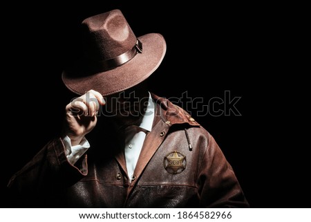 Photo of a shaded sheriff officer with badge in jacket putting on cowboy hat on black background. Royalty-Free Stock Photo #1864582966