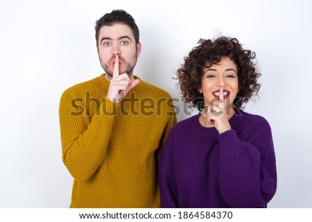 Smiling Young couple wearing knitted sweater standing against white background makes shush gesture, holds fore finger over lips hides secret. Be mute, please.