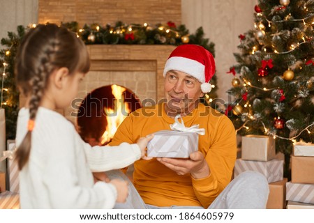 Grandfather with granddaughter giving present on Christmas, child with pigtails in white jumper congratulating her grandfather, senior man in santa hat and yellow casual sweater.