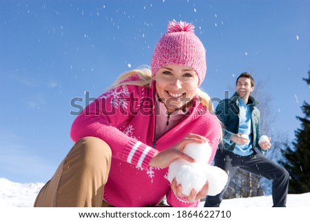 Young woman crouching in a field of snow and holding snow balls while smiling at the camera with her husband in the background