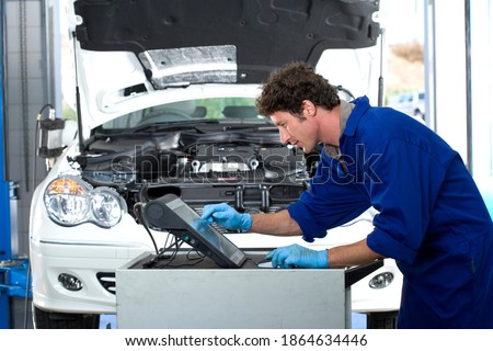 Middle-aged car mechanic wearing blue overalls and using a computer in and auto repair shop with a broken down car in the background