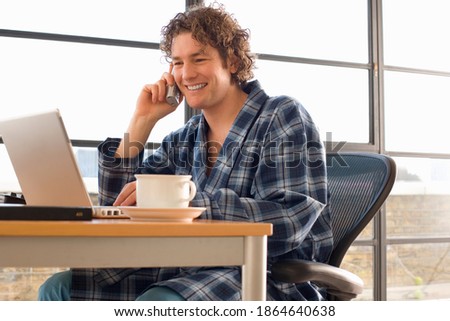 Young man sitting at his desk in his home office and talking on the telephone while wearing a dressing gown and smiling Royalty-Free Stock Photo #1864640638