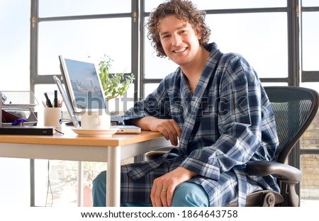 Young man sitting at his desk in his home office and leaning on the table while wearing a dressing gown and smiling Royalty-Free Stock Photo #1864643758