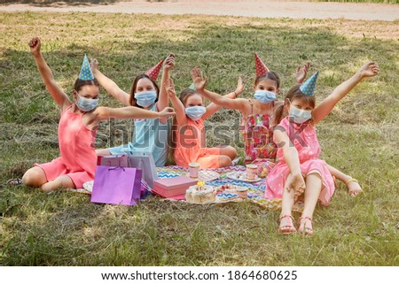 Birthday party in medical masks during the pandemic. Children's birthday celebration in a medical mask. Girls in bright dresses sit on a blanket on the grass.