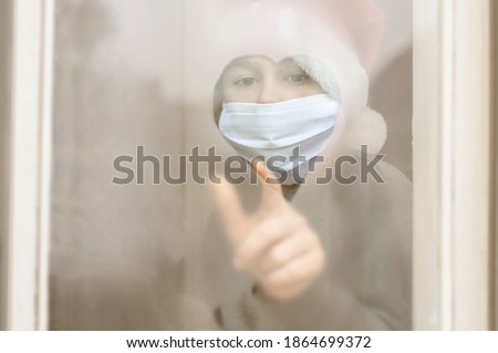 Art noise. Child in a santa claus hat, protective mask draws a heart on the foggy misted window with his finger. Christmas holidays 2021. Covid-19, Coronavirus. Waiting. Isolation at home. Hospital