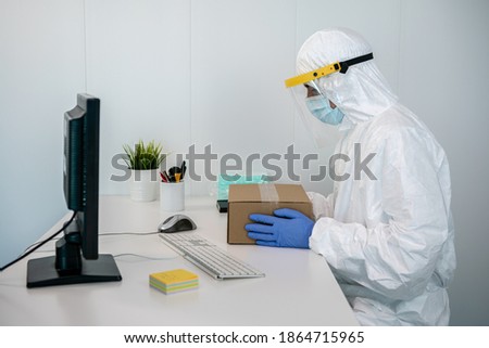 Doctor in protective suit and plastic face shield packet new medical masks in a box during Covid19 at hospital. Man wearing hand gloves and PPE in interior of clinic setup amid Coronavirus Pandemic.