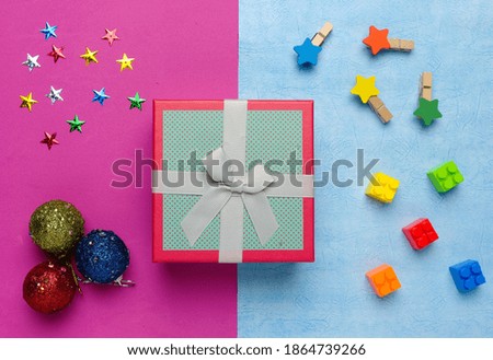 Flatlay picture of gift box with building block, stars, Christmas ball and stars on pink and blue background.