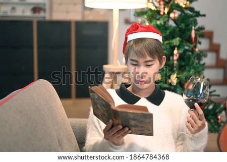 A young woman holding wine glass and reading book with Christmas tree in background.
