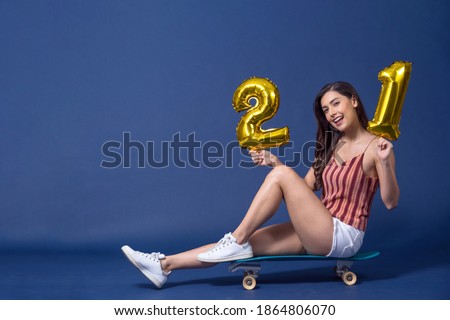 Happy young Asian woman sitting on surfskate or skateboard and holding 21 gold color balloons for celebrate merry Christmas and happy new year on blue color background, exercise and sport concept