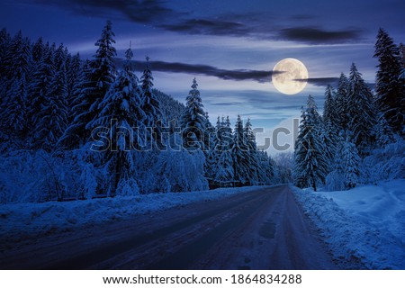 road through mountain landscape in winter at night. spruce forest covered in snow. dramatic sky with clouds glowing in full moon light