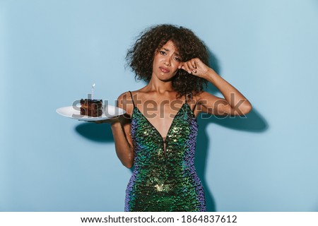Sad charming woman with fluffy curly hairstyle in shiny modern dress looking into camera and holding piece of cake with candle on blue backdrop..