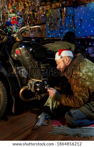 A man in a santa claus hat repairs an old motorcycle in the garage on New Year's Eve.