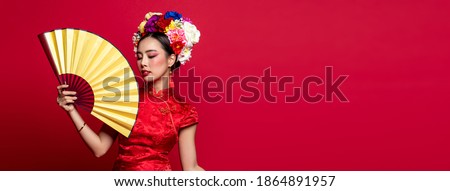Beautiful Asian woman wearing traditional cheongsam qipao dress holding golden fan in isolated colorful red banner background with copy space