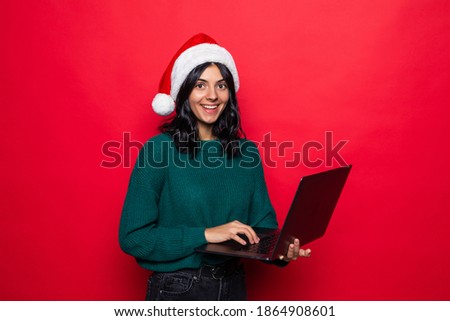 Attractive woman with santa's christmas hat uses laptop on red background.