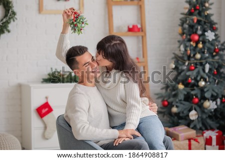 Young woman kissing her husband under mistletoe branch at home on Christmas eve