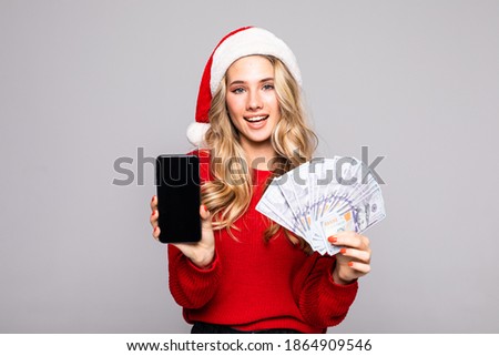 Portrait of a young successful woman in red christmas hat holding bunch of money banknotes while using mobile phone over white background
