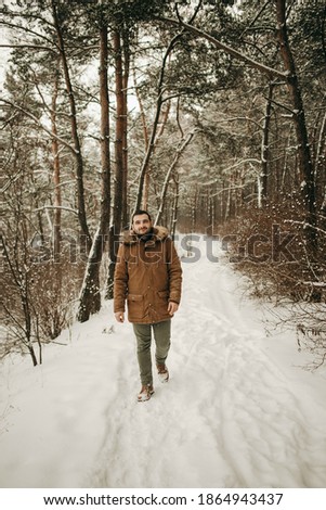 Portrait of a young handsome European man walking in a brown jacket in a snowy pine forest. Park, snowfall, trees, loneliness, travel, winter, walk. Beautiful guy.