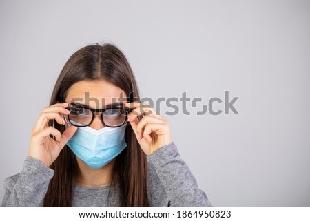 Woman wiping foggy glasses caused by wearing medical mask indoors, closeup. Woman wiping foggy glasses caused by wearing disposable mask on gray background, closeup.