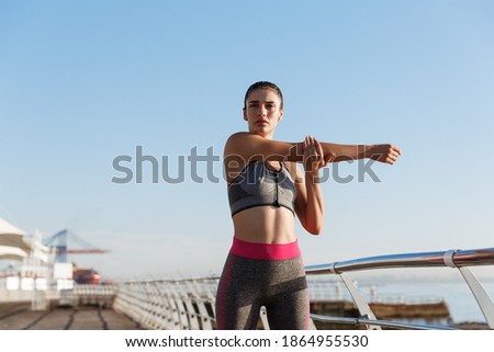 Image of beautiful healthy woman in fitness clothing stretching her body before jogging training. Female runner standing on the seaside promenade, training near the sea