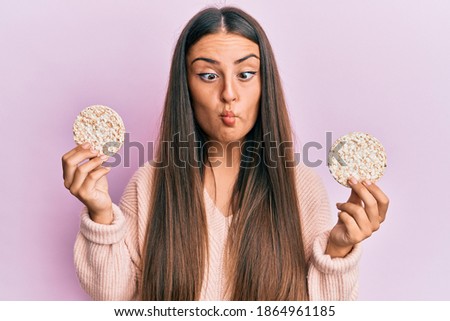 Beautiful hispanic woman eating healthy rice crackers making fish face with mouth and squinting eyes, crazy and comical. 