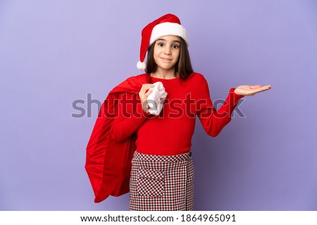 Little girl with hat and Christmas sack isolated on purple background having doubts while raising hands