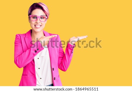 Young beautiful woman with pink hair wearing business jacket and glasses amazed and smiling to the camera while presenting with hand and pointing with finger. 