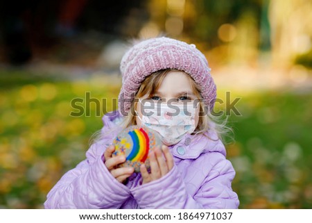 Adorable little toddler girl with medical mask and painted rainbow on stone during pandemic coronavirus quarantine. Cute smiling child outdoors. Rainbows as hope, sign and fight against corona virus