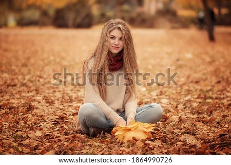 Beautiful girl sitting on the leaves in the autumn forest. She has yellow leaves in her hands. Image with selective focus and toning. Image with noise effects. Focus on the eyes.