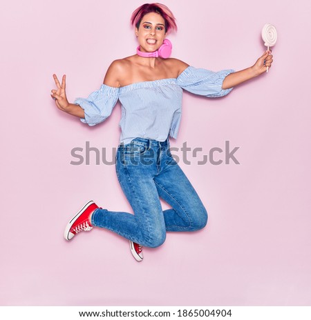 Young beautiful woman with pink short hair doing victory sign smiling happy. Jumping with smile on face using headphones holding lollipop over isolated background.