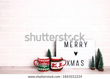 Merry Xmas text on white Lightbox with miniature Christmas trees and a couple of mugs against white brick wall. Copy space for text.